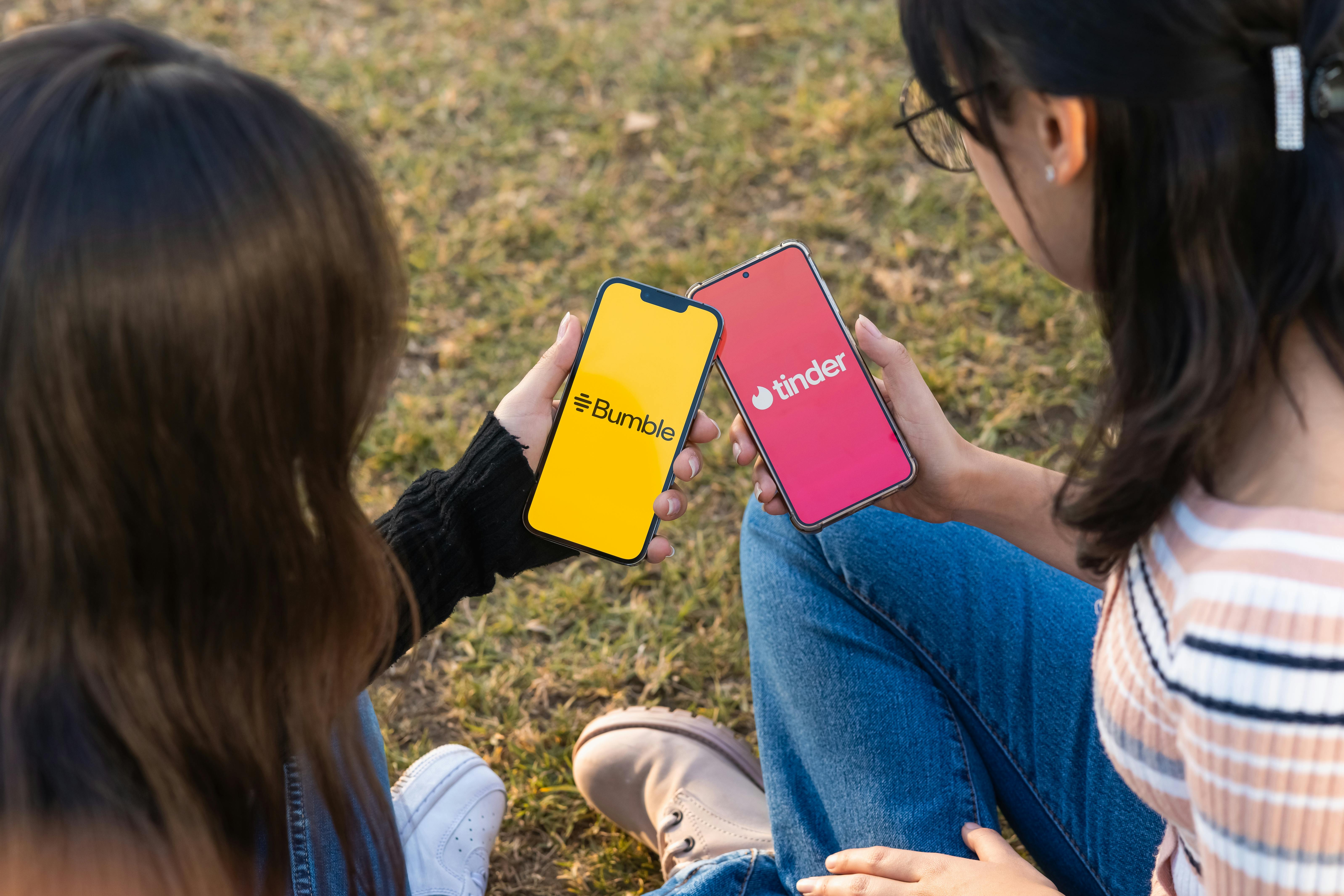 Two women holding phones open to the "Bumble" and "Tinder" dating apps.